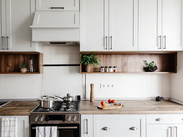 Cooking food in modern, simple kitchen with white cabinets and wood tabletop. Knife on wooden cutting board with vegetables, pepper, spices.