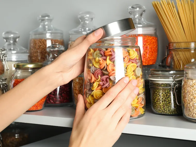Woman cooking from pantry taking jar of colorful pasta from white shelf, closeup
